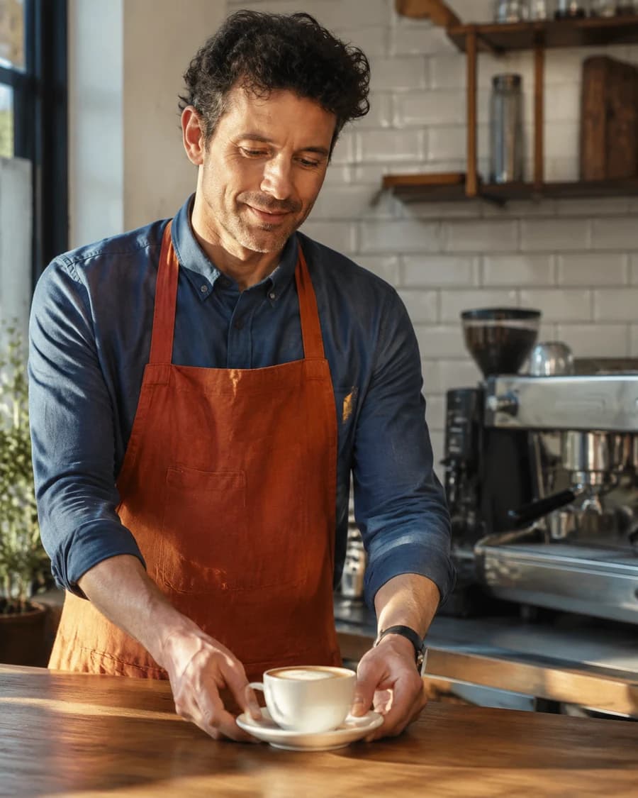 A café owner in a rust apron at his coffee counter